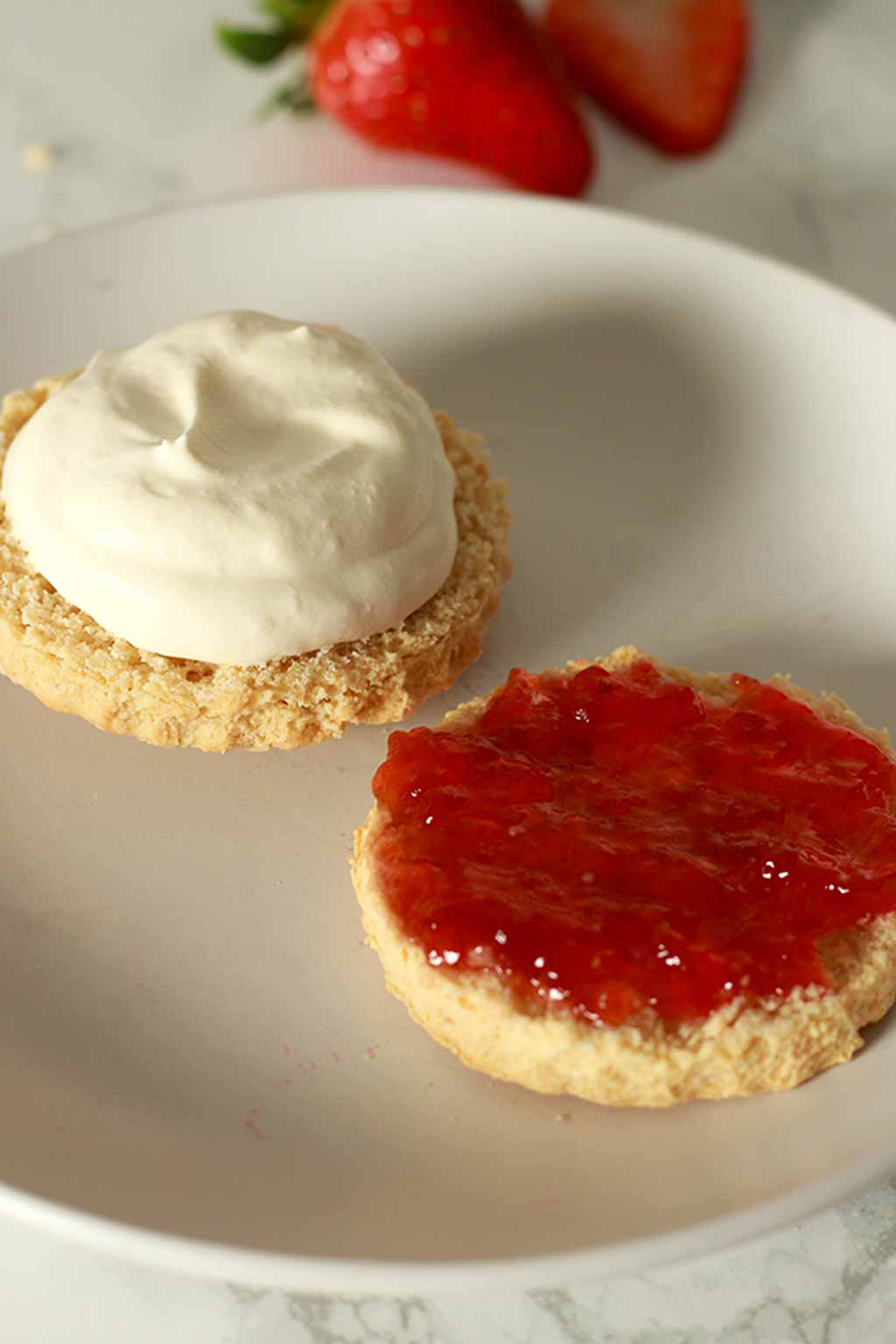 Two Halves Of A Scone On A White Plate. One Has Jam On It And The Other Has Cream On It.