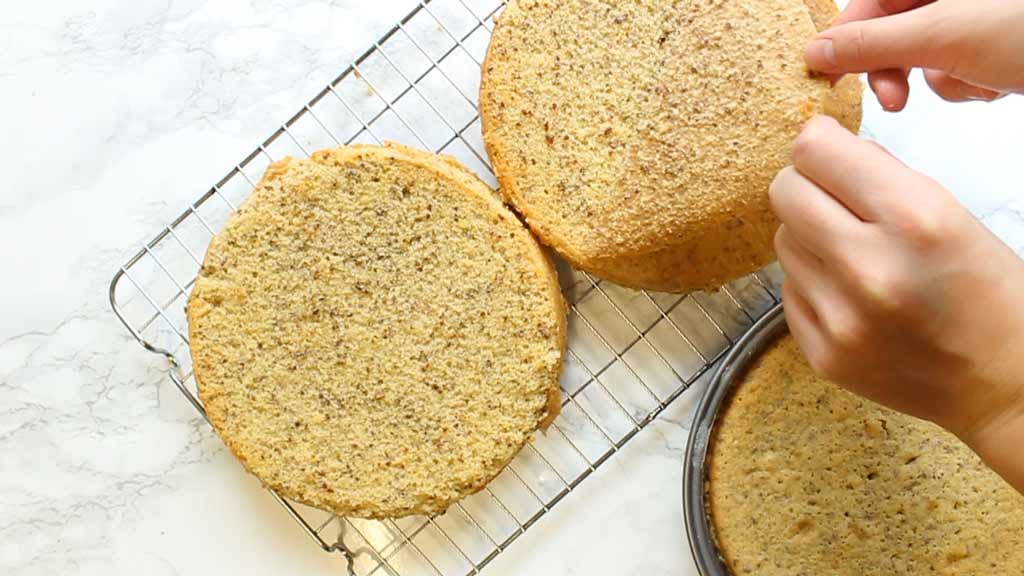 Two baked cakes on a cooling rack. Person is peeling off parchment paper from the bottom of one of the cakes.