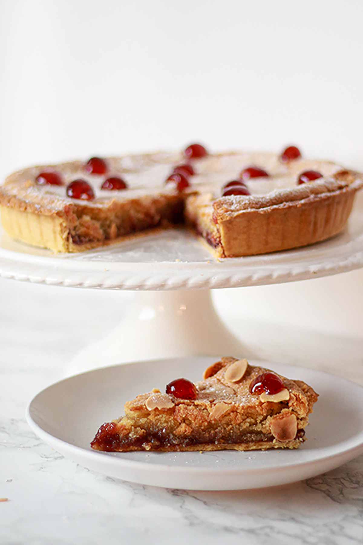 Slice Of Cherry Bakewell Tart With The Rest Behind It On A White Cake Stand