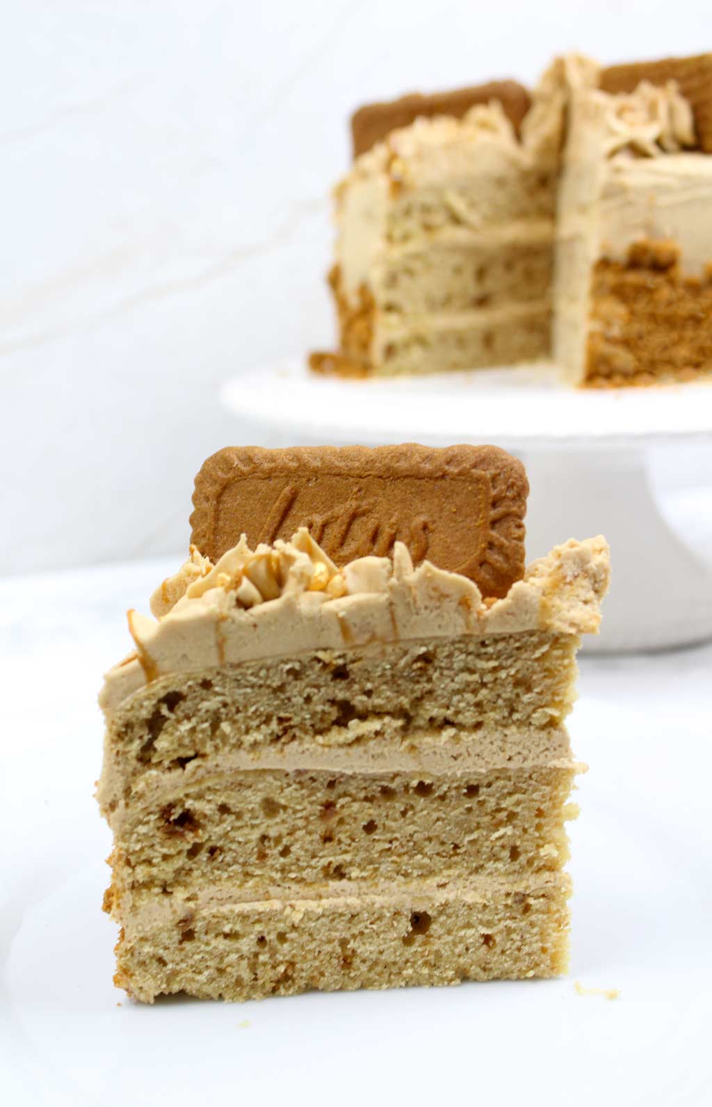 a slice of biscoff cake with the rest of the cake behind it on a cake stand