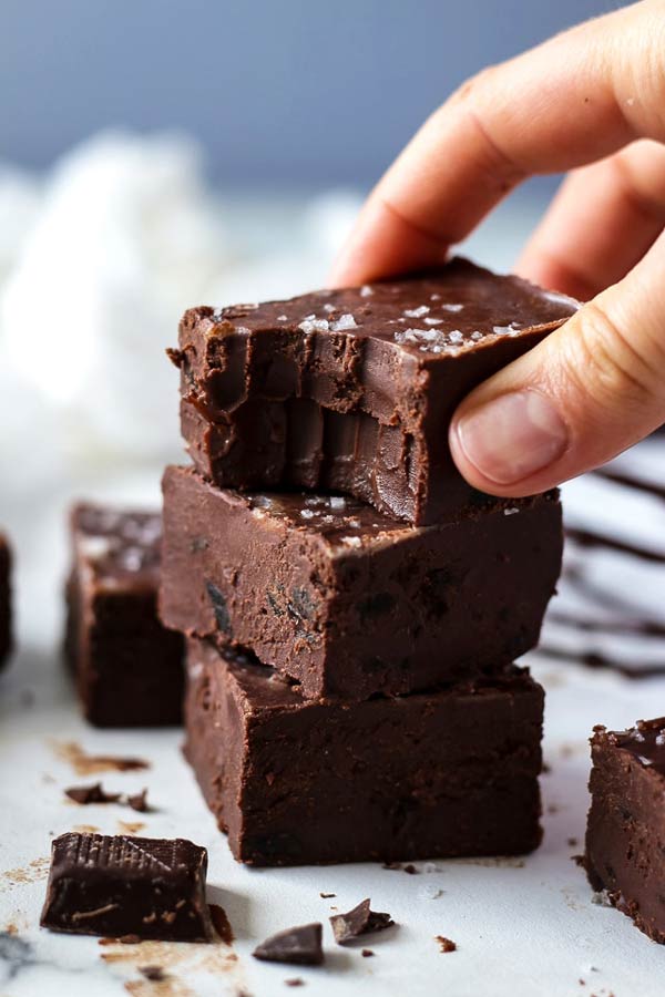 3 pieces of chocolate fudge stacked on top of eachother. The top piece has been bitten out of and a hand is reaching for it.