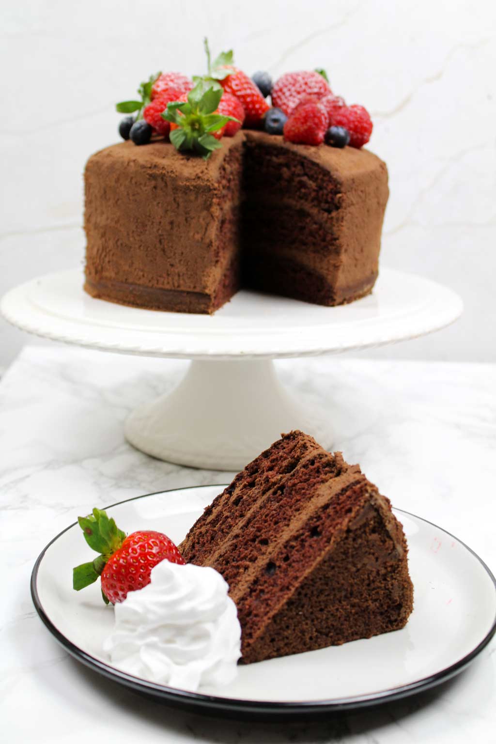 slice of vegan chocolate cake on a plate with whipped cream and a strawberry beside it. The rest of the cake is on a cake stand in the background