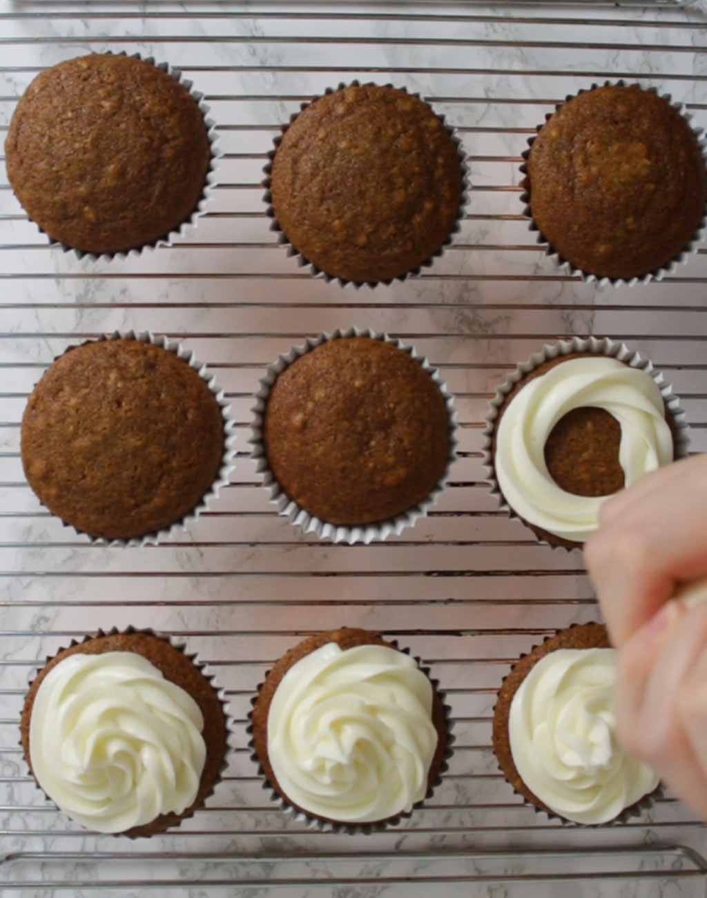 Piping Cream Cheese Frosting Onto A Cupcake Sitting On A Wire Rack