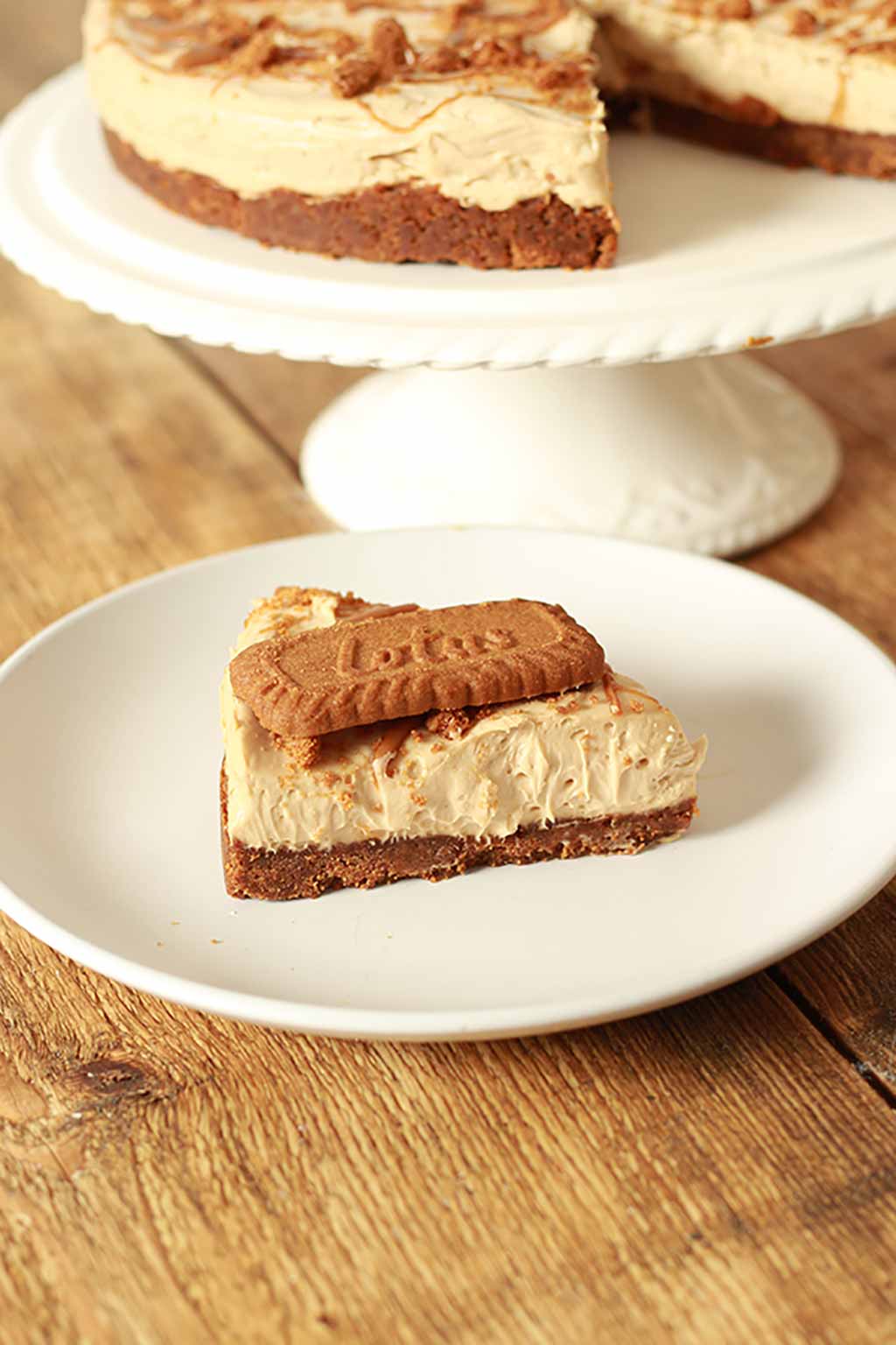 Slice Of vegan Biscoff Cheesecake On A White Plate With Remaining Cheesecake In The Background